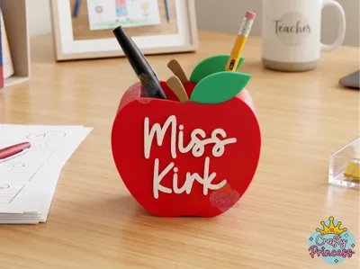 Red 3D-printed apple-shaped pencil holder with the name Miss Kirk embossed in white, filled with pens on a teacher's desk